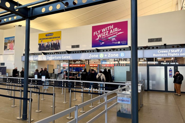 Airport departure area with people in line and flight advertisements above.