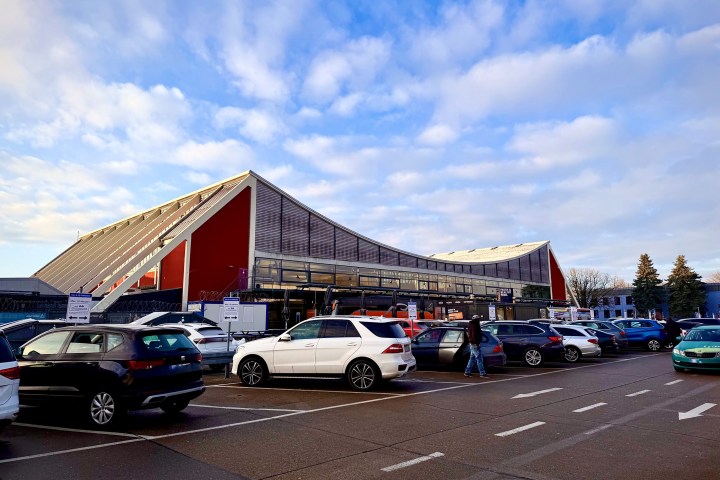 Modern building with sloped roof and parked cars in foreground under a cloudy sky.