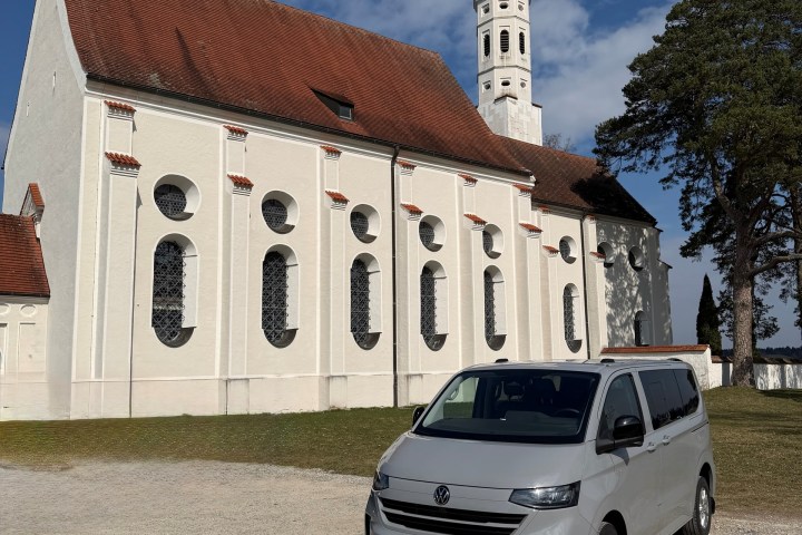 Gray van parked near white church with tall tower under a blue sky.
